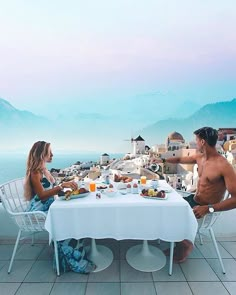 a man and woman sitting at a table with food on it in front of the ocean