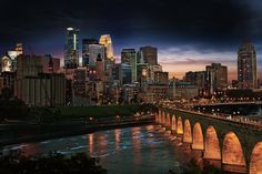 the city skyline is lit up at night with lights on and bridge over water in foreground