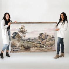 two women standing next to each other holding up a painting with sheep in the background