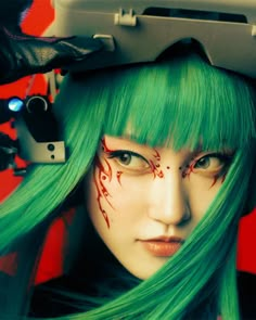 a woman with green hair and makeup has her head painted like a demon, holding a video game controller over her head