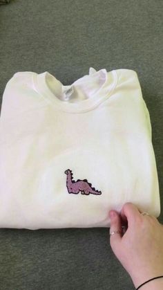 a person holding onto a white t - shirt with a pink dinosaur embroidered on it