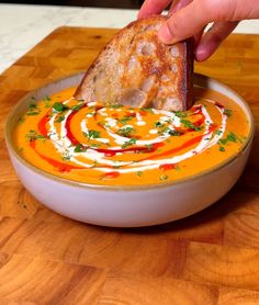 a hand dipping a piece of bread into a bowl of carrot soup with red sauce