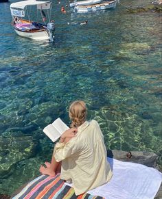 a woman reading a book while sitting on a towel in the water next to boats