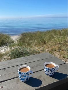 two cups of coffee sitting on top of a wooden table next to the ocean and grass