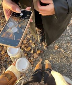 two people sitting on the ground taking pictures with their cell phones and coffee cups in front of them