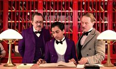 three men in tuxedos stand at a desk with an open book and pen