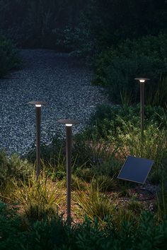 three solar powered lights in the middle of some grass and bushes, with a gravel area behind them