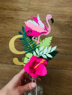 a person holding a vase with flowers and paper flamingos in it on a wooden table