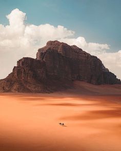 two people riding camels in the desert with mountains and clouds behind them on a sunny day