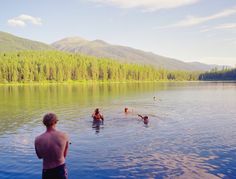 some people are swimming in the water with mountains in the backgrouds and trees