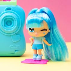 a doll with blue hair standing next to a small camera and toy on a table