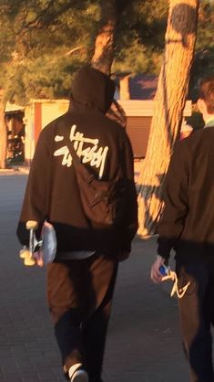 two men walking down the street with skateboards in their hands and one man wearing a black hoodie