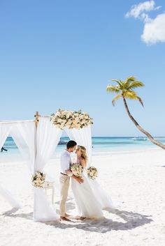 a bride and groom standing under an arch on the beach with palm trees in the background