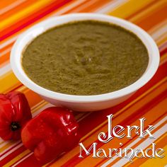 two red peppers sitting next to a white bowl filled with green pesto sauce on a striped tablecloth