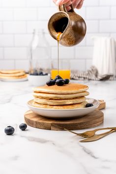 a person pouring syrup on top of pancakes with blueberries and oranges in the background