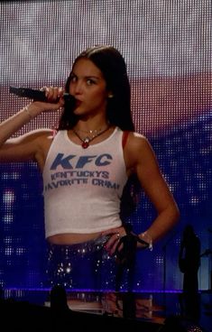 a woman standing on stage holding a microphone in her right hand and wearing a white tank top