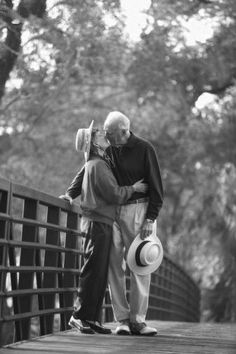 an older man and young woman kissing on a bridge with trees in the back ground