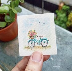 a person holding up a card with a bicycle and flowers on the front, next to a potted plant