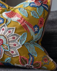 a yellow pillow with blue and red flowers on it sitting on a gray couch next to a window