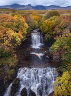 an aerial view of a waterfall surrounded by trees in the fall with mountains in the background