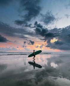 a person holding a surfboard walking on the beach at sunset with clouds in the background