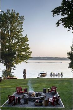 a fire pit in the middle of a grassy area with chairs around it and a lake in the background