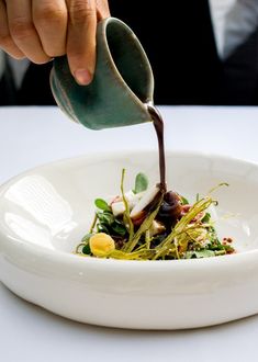 a person pouring sauce on top of a white bowl filled with vegetables and other things