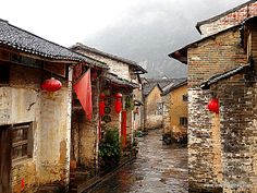 an alley way with stone buildings and red lanterns hanging from the roof, on a rainy day