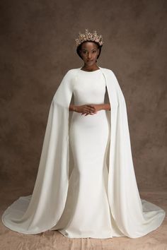 a woman wearing a white gown and cape with a crown on her head, standing in front of a brown background