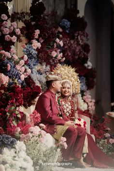 a man and woman dressed in traditional thai garb sitting on a bench surrounded by flowers