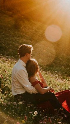 a man and woman sitting in the grass with their arms around each other, looking into the distance