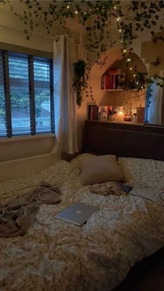 an unmade bed with a laptop on it in front of a window and bookshelf