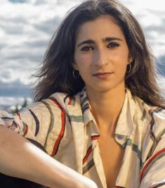 a woman with long hair sitting in front of the ocean wearing a shirt and earrings