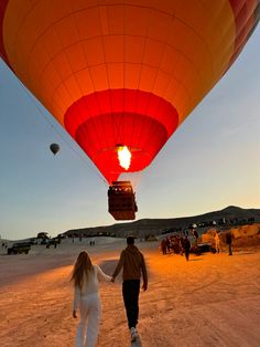 a man and woman holding hands while walking in front of a hot air balloon