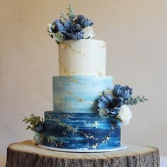 a three tiered blue and white cake with flowers on top is sitting on a tree stump