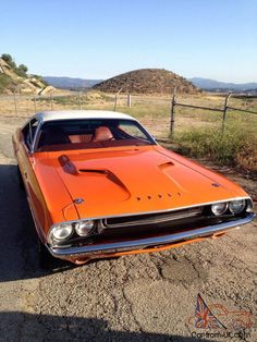 an orange muscle car parked on the side of a dirt road next to a fence