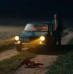 a man standing next to a car on a dirt road at night with the lights on