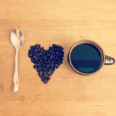 a cup of coffee next to a spoon and heart shaped beans on a wooden table