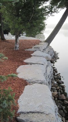 a stone path next to a body of water with trees on both sides and chairs in the background