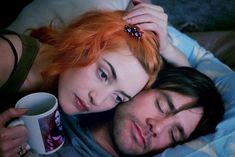 a man and woman laying next to each other on top of a bed holding coffee mugs