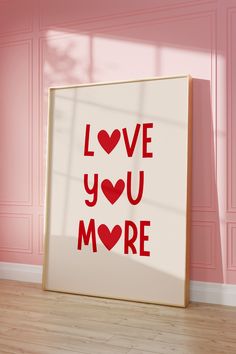 a framed poster with the words love you more on it in front of a pink wall