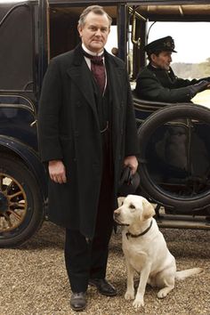 a man standing next to a white dog in front of an old fashioned car with two men