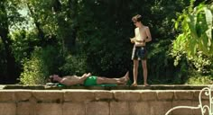 a man standing next to a woman laying on top of a stone wall near trees