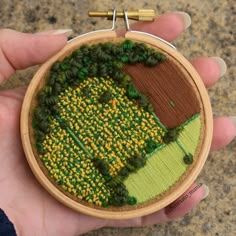 a hand holding a small embroidery hoop with trees and yellow flowers on the outside, in front of a stone wall