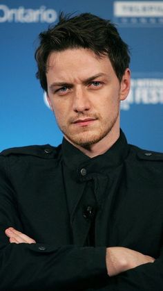 a man in black shirt and tie standing with his arms crossed looking at the camera