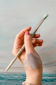 a person holding a paintbrush and some white powder on their face with the ocean in the background