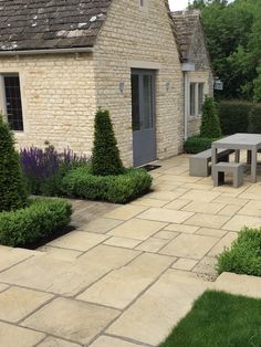 an outdoor patio with table and benches surrounded by bushes, shrubs and trees in front of a stone building