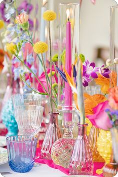 colorful vases and candles on a table with flowers in the center, surrounded by other glassware