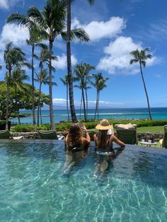 two women sitting on the edge of a swimming pool with palm trees in the background