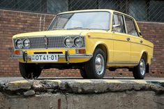 an old yellow car parked on the side of a road next to a brick wall